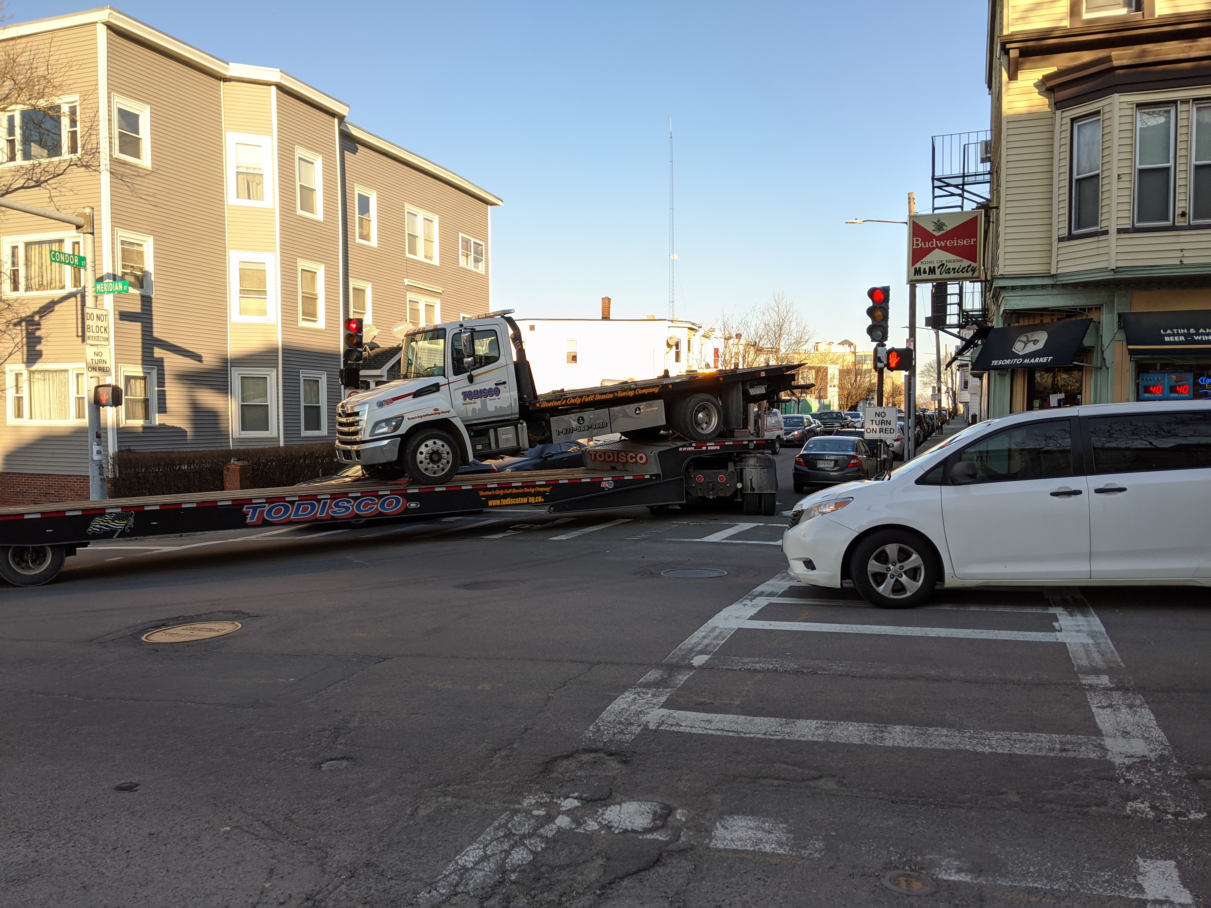 A semit truck attempts a tight turn in an urban area. There are buildings on each side of the street, along with parked cars, and the streets are narrow. The truck swung wide past the parked cars and is now turning hard to get back into the travel lane.